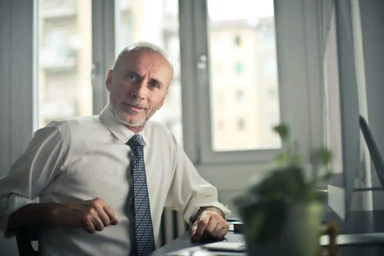 Professional middle-aged man in white shirt and patterned tie sitting at desk by window.