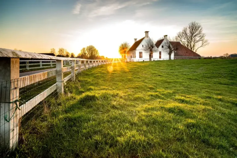 Sunset over green grassy field with white wooden fence and traditional white farmhouse.