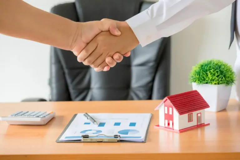 Two people shaking hands over a desk with documents, a small house model, and a potted plant.