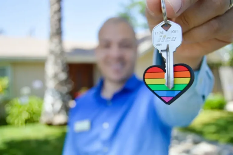 Person holding a silver key with a rainbow-striped heart keychain outside a residential house.