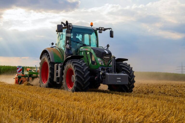 Green and black tractor with red wheels plowing a golden field under cloudy sky.