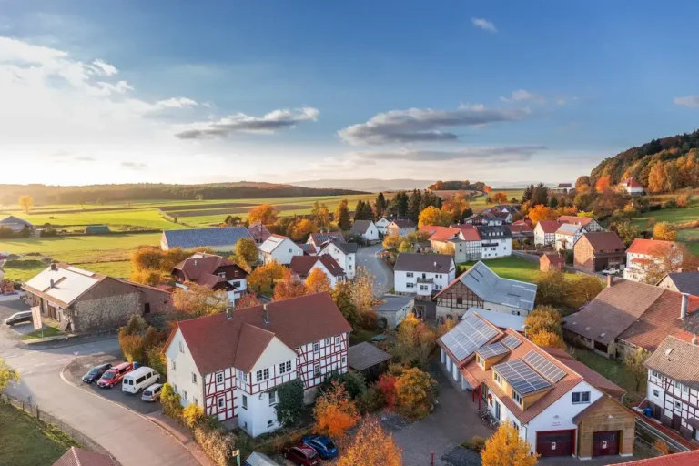 Aerial view of a rural village with red-roofed houses, solar panels, autumn trees, and green fields under a blue sky.