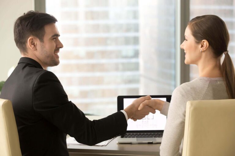 Two professionals, a man in a black suit and a woman in a light sweater, shaking hands in an office setting.