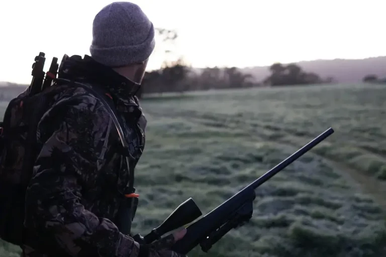 Man in camouflage clothing and gray beanie holding a rifle in a grassy field at dawn.