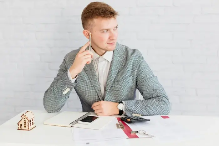 Young professional man in gray blazer sitting at desk with house model, documents, and calculator.