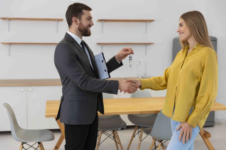 A man in a suit handing keys to a woman in a yellow blouse while shaking hands.