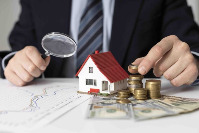 Person in suit examining model house with magnifying glass and stacking coins on US dollar bills.