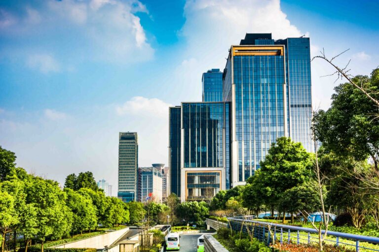 Urban scene with modern glass office buildings, green trees, and a clear blue sky.
