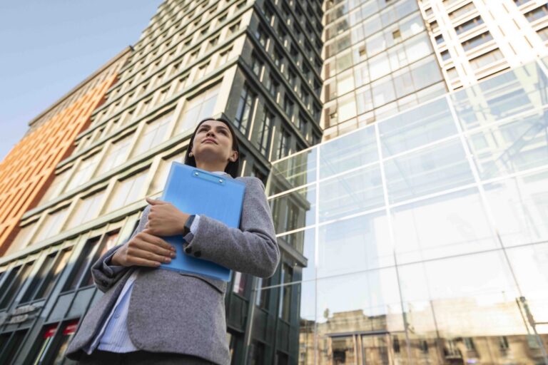 Professional woman holding blue clipboard standing in front of modern office buildings.