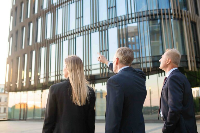 Three professionally dressed individuals observing and discussing a modern glass building facade at sunset.