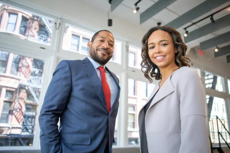 Two professionally dressed businesspeople, a man in a blue suit with a red tie and a woman in a light gray blazer, standing in a modern office with large windows.