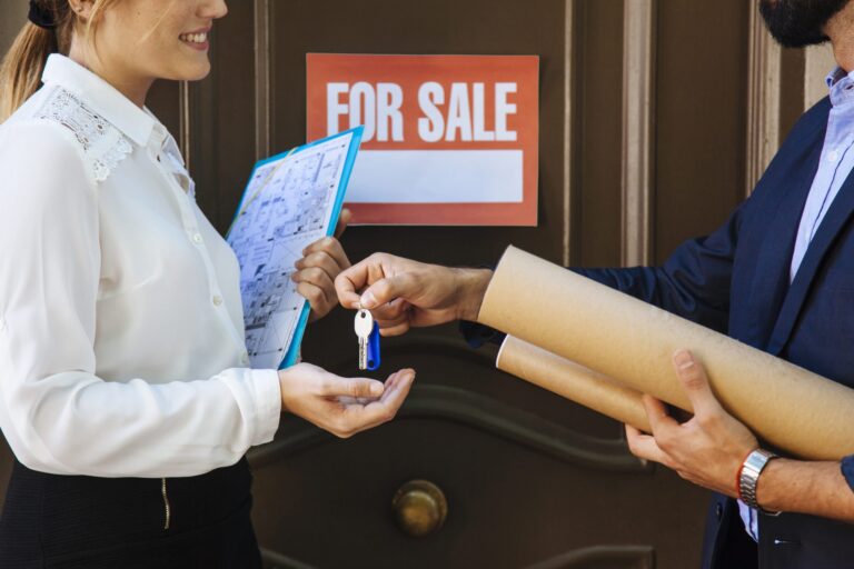 Two professionals exchanging house keys in front of a door with a "For Sale" sign.