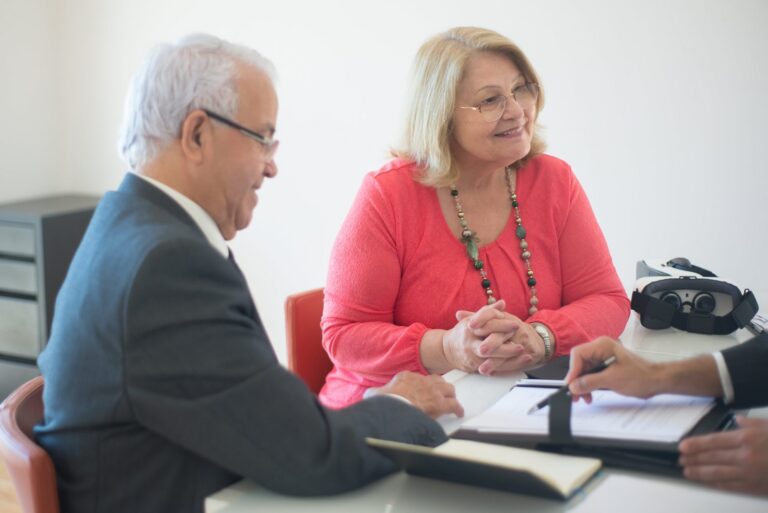 Two elderly professionals in discussion, one woman in coral blouse and man in suit, with documents on table.