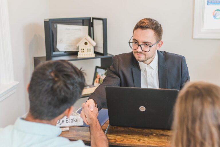 Mortgage broker in suit shaking hands with clients across wooden desk in office.