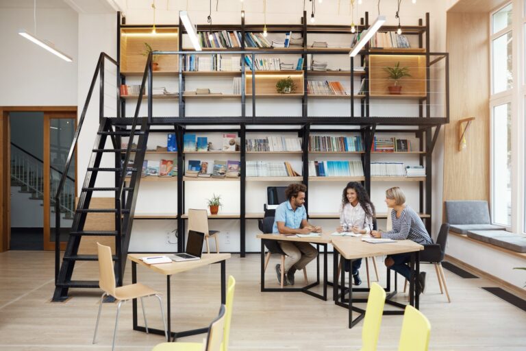Three people engaged in discussion at a modern library with black metal shelves and wooden tables.