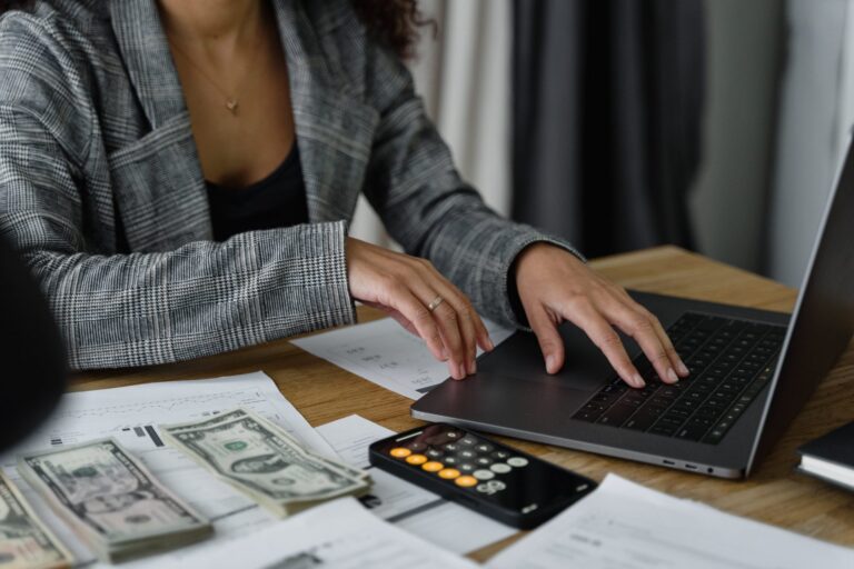 Person in a gray plaid blazer working on a laptop with cash, calculator, and documents on wooden desk.