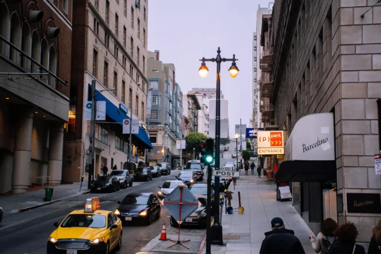 Urban street scene at dusk with traffic, pedestrians, historic buildings, and illuminated signage.