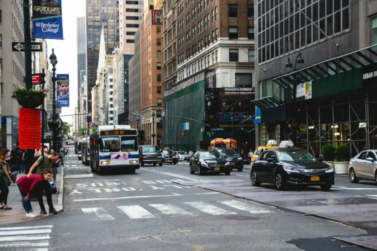 Busy urban street with buses, cars, pedestrians, and tall buildings under cloudy sky.