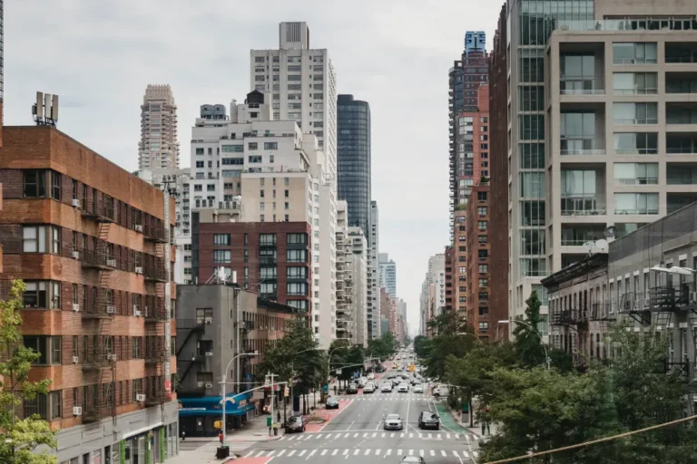 Urban street view with mid-rise and high-rise buildings, cars, and trees on a cloudy day.