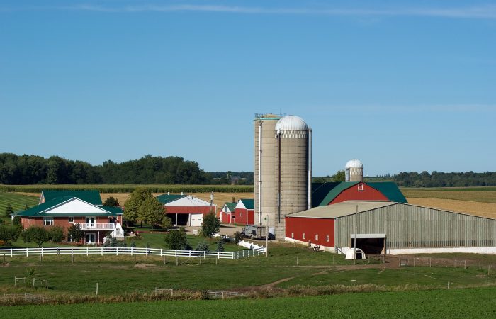 Rural farmstead with red barns, silos, green roofs, white fencing, and surrounding fields under clear blue sky.