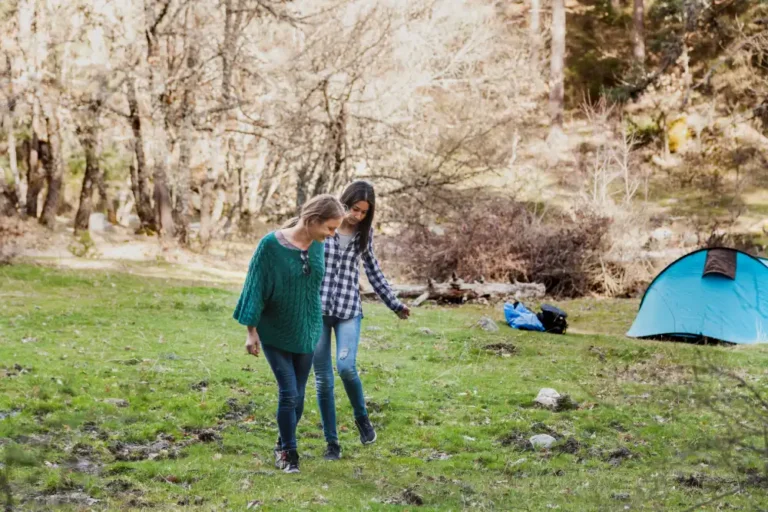 Two women walking on grassy terrain near a blue camping tent in a wooded area.