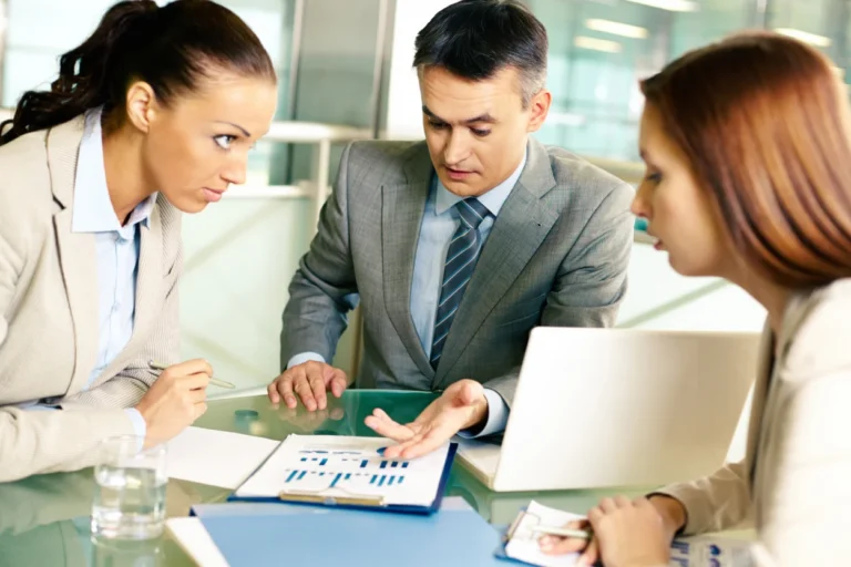 Three business professionals in formal attire discussing charts and documents at a glass table.