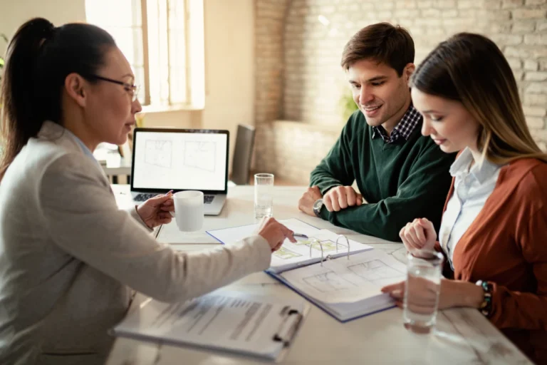 Three professionals reviewing architectural plans at a table with a laptop and beverages.