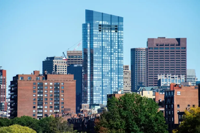 City skyline featuring modern glass skyscraper and brick buildings with green trees in foreground.