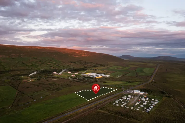 Aerial view of a rural landscape with green fields, scattered buildings, vehicles, and a highlighted plot outlined in white with a red house icon.