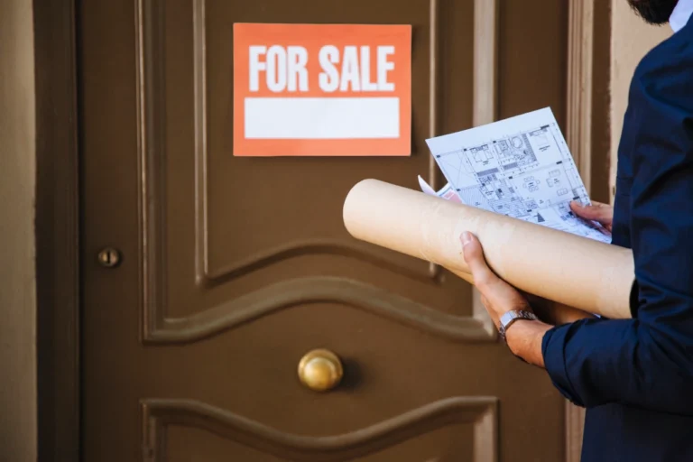Person holding architectural blueprints near brown door with orange "For Sale" sign.