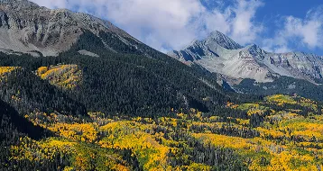 Mountain landscape with green and yellow autumn trees under a partly cloudy sky.