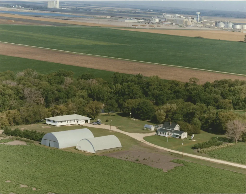 Aerial view of a rural farm with green fields, trees, two white storage buildings, and two houses.