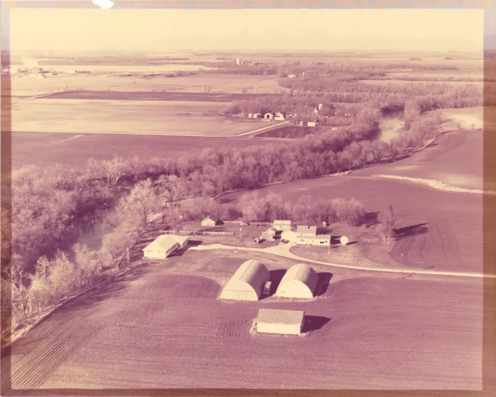 Aerial view of a rural farmstead with buildings, silos, a winding river, and surrounding farmland.