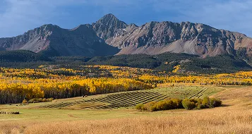 Panoramic view of rugged mountains with yellow autumn trees and grassy fields in foreground.