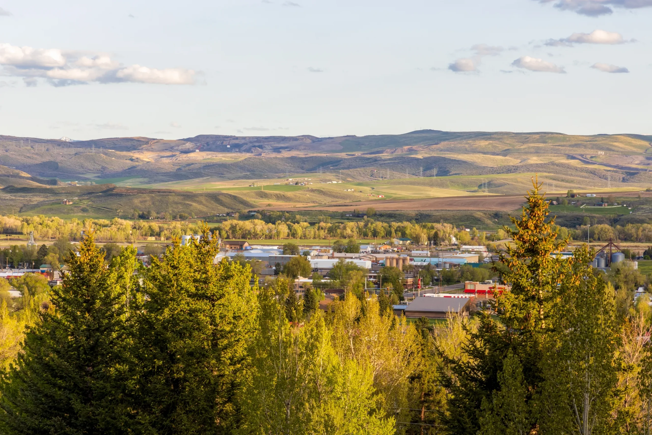 Panoramic view of a small town surrounded by green trees and rolling hills under a clear sky.
