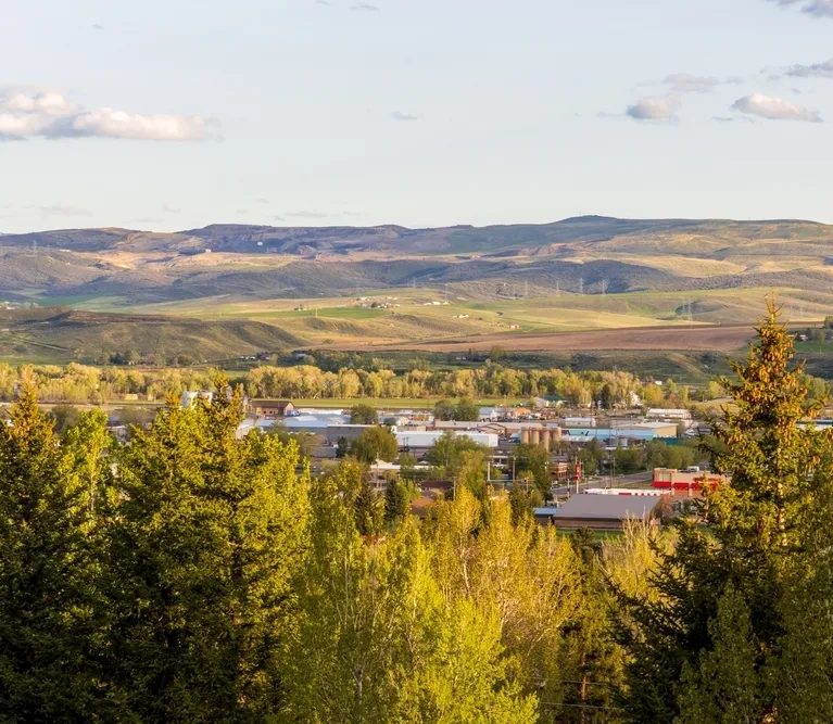 Panoramic view of a small town surrounded by green trees with rolling hills and farmland in the background under a partly cloudy sky.