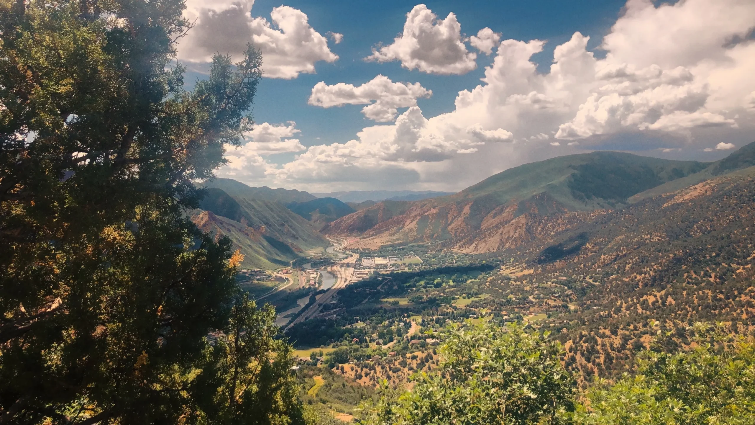 Scenic valley view with green trees, winding river, mountainous terrain, and partly cloudy sky.