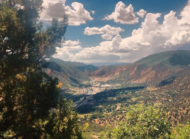 Panoramic view of a valley town surrounded by green and brown mountains under a partly cloudy blue sky.
