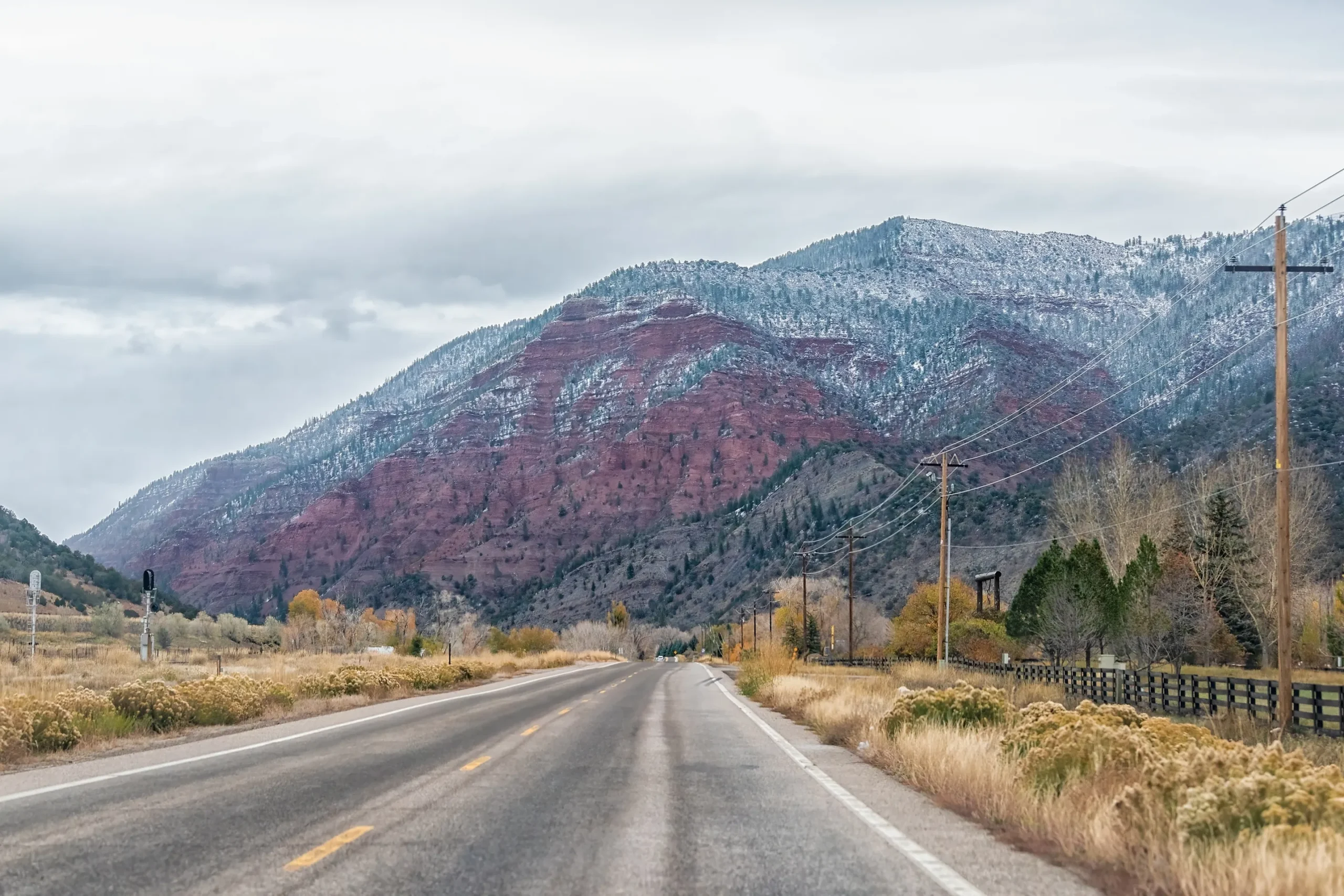 Two-lane road leading toward red rock mountains with light snow and overcast sky.