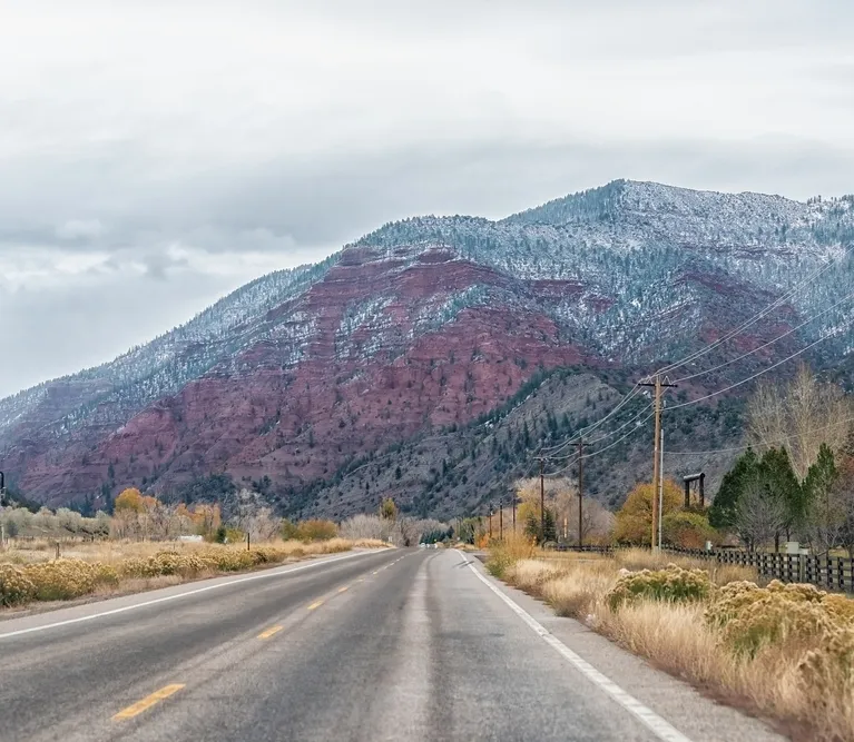 Two-lane road leading toward red rock mountains with light snow and overcast sky.