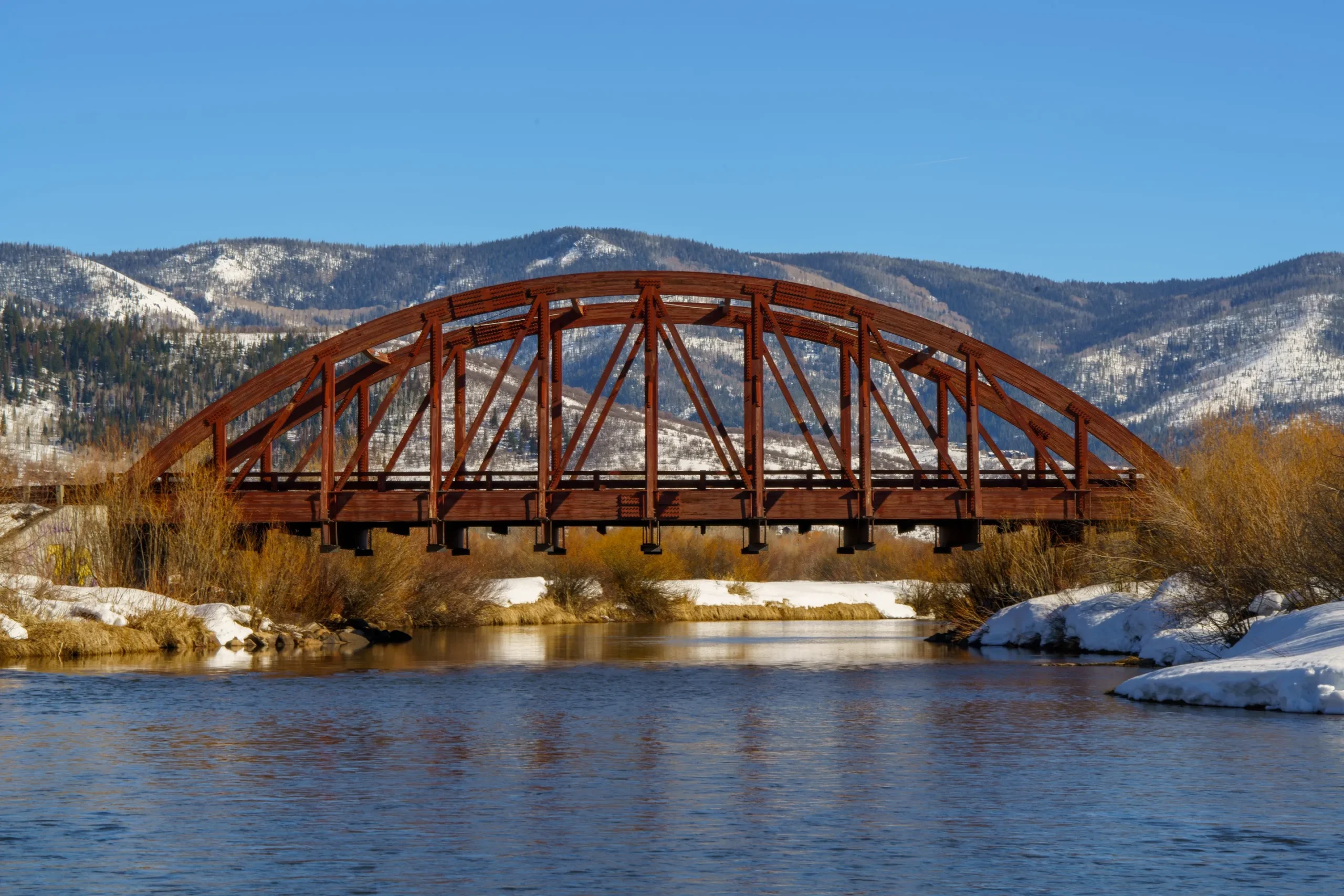 Rust-colored steel arch bridge spanning a calm river with snow-covered banks and mountainous background.