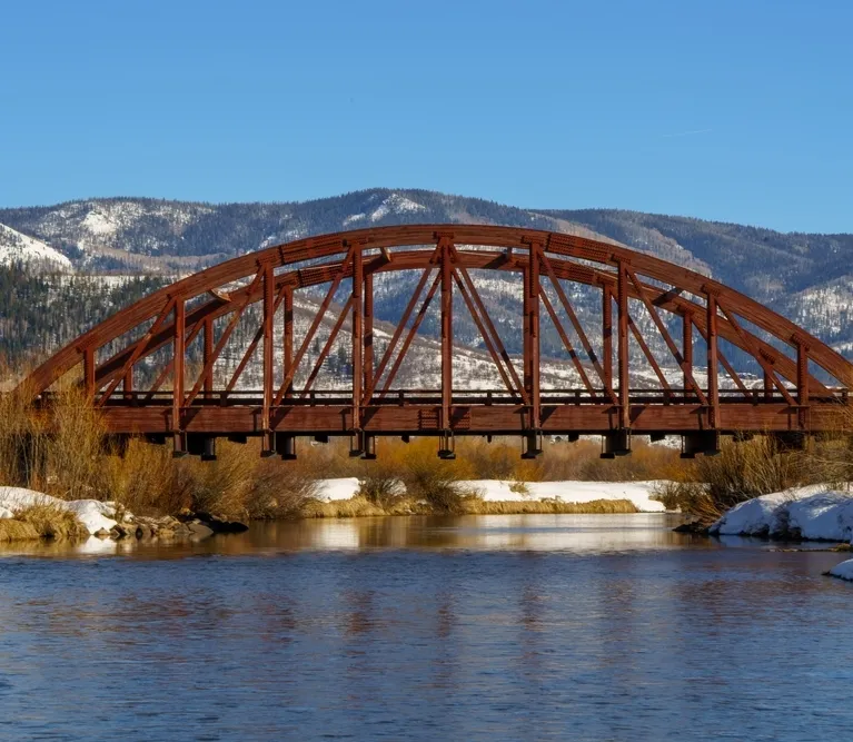 Rust-colored metal arch bridge spanning a calm river with snow-covered banks and forested mountains in the background.