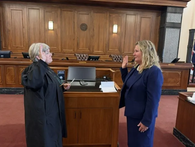 Two women in a courtroom, one administering an oath to the other, both with raised right hands.