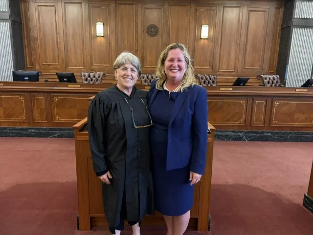 Two women standing in a courtroom, one in judicial robes and the other in a navy blue business suit.
