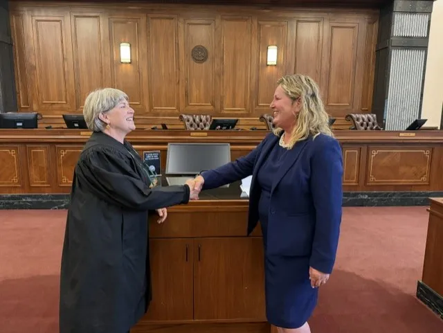 Two women shaking hands in a courtroom, one in a judge's robe, the other in a navy suit.