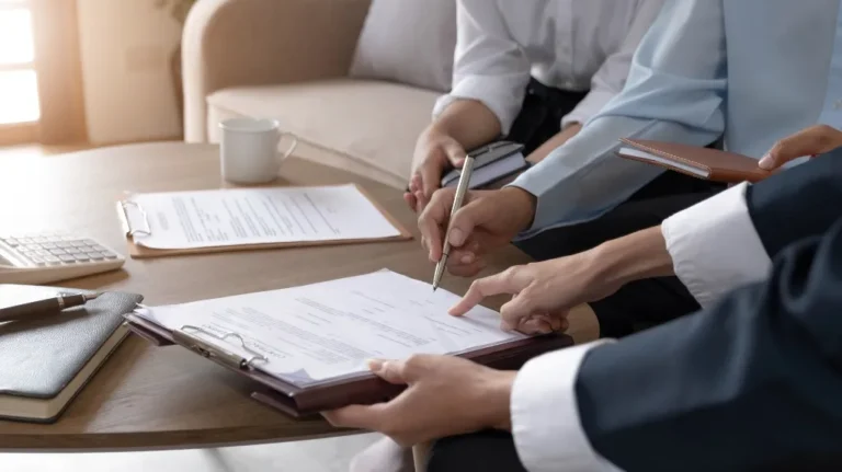 Three people in formal attire reviewing and signing documents on a wooden table.