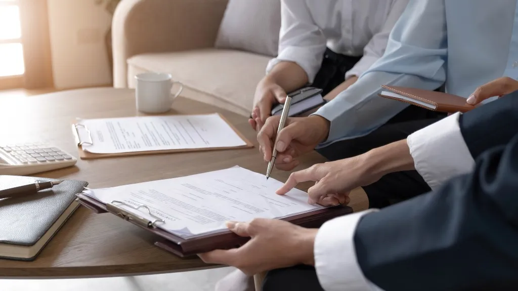 Three people in formal attire reviewing and signing documents on a wooden table.