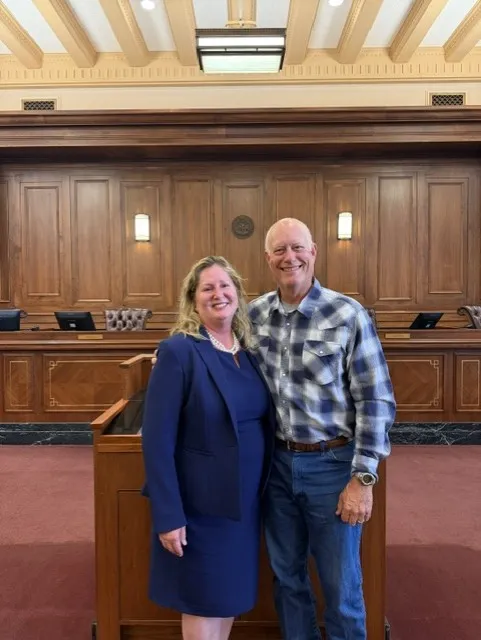 Two individuals, a woman in a navy blue suit and a man in a plaid shirt and jeans, standing and smiling inside a formal courtroom with wooden paneling.