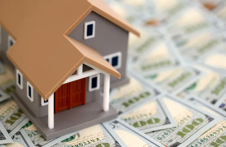 Small gray and brown model house placed on scattered US hundred-dollar bills.