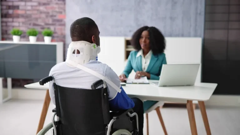 Man in a wheelchair wearing a neck brace, consulting with a professional woman in an office.